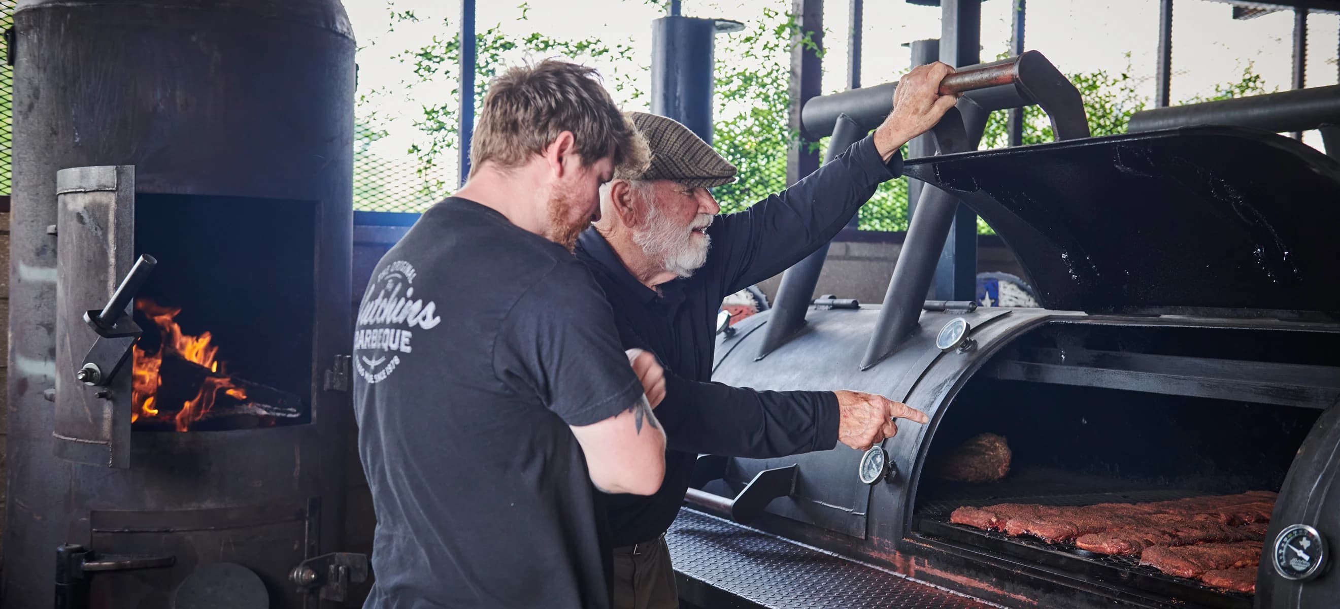 Two men stand near a large BBQ smoker and inspect the contents