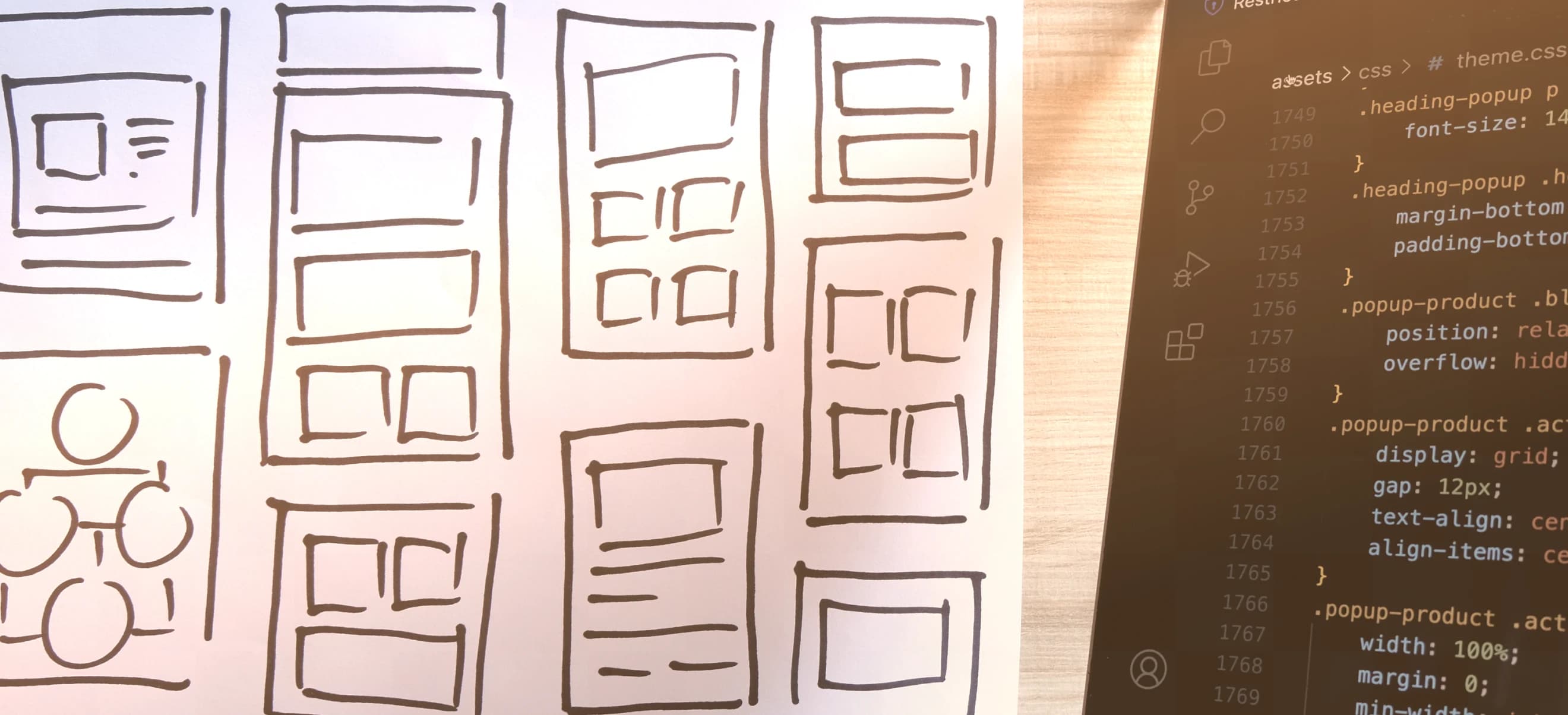 A workspace with hand-drawn sketches of website layouts on a piece of paper, next to a laptop screen displaying CSS code.