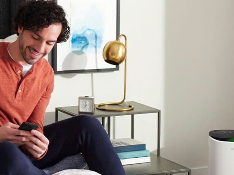 A man sits on a bed using a 3M Filtrete app, with a bedside table and an air purifier next to him,