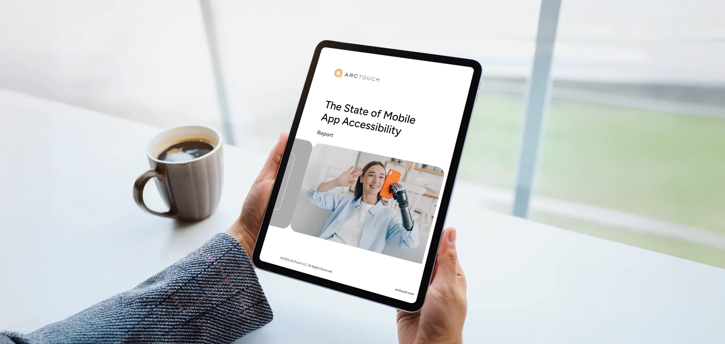 Two hands holding a tablet over a white table with a coffee cup on the left. On the tablet screen is the cover of the ArcTouch State of Mobile App Accessibility report.