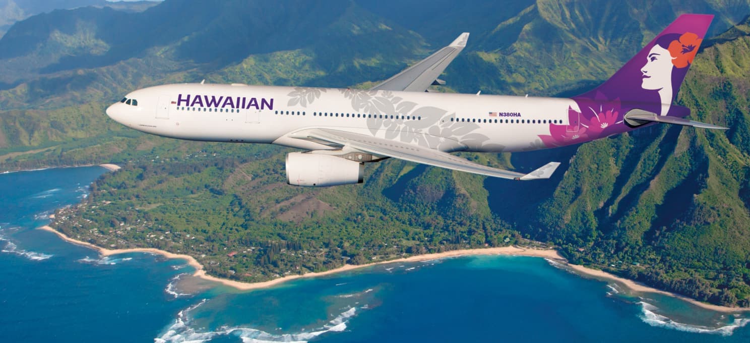 A Hawaiian Airlines airplane flies over a scenic coastline with lush green mountains and blue ocean waters