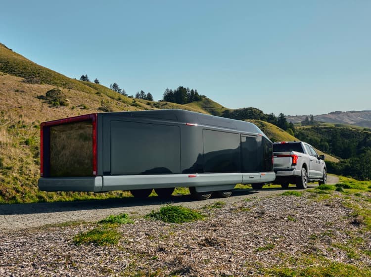 A truck tows a streamlined, modern Lightship along a scenic, hilly road, reminiscent of a picturesque case study in efficient transportation.