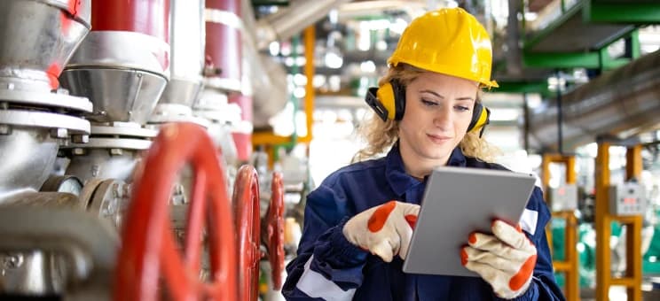 A woman dressed in protective work clothing operates a tablet in an industrial environment.