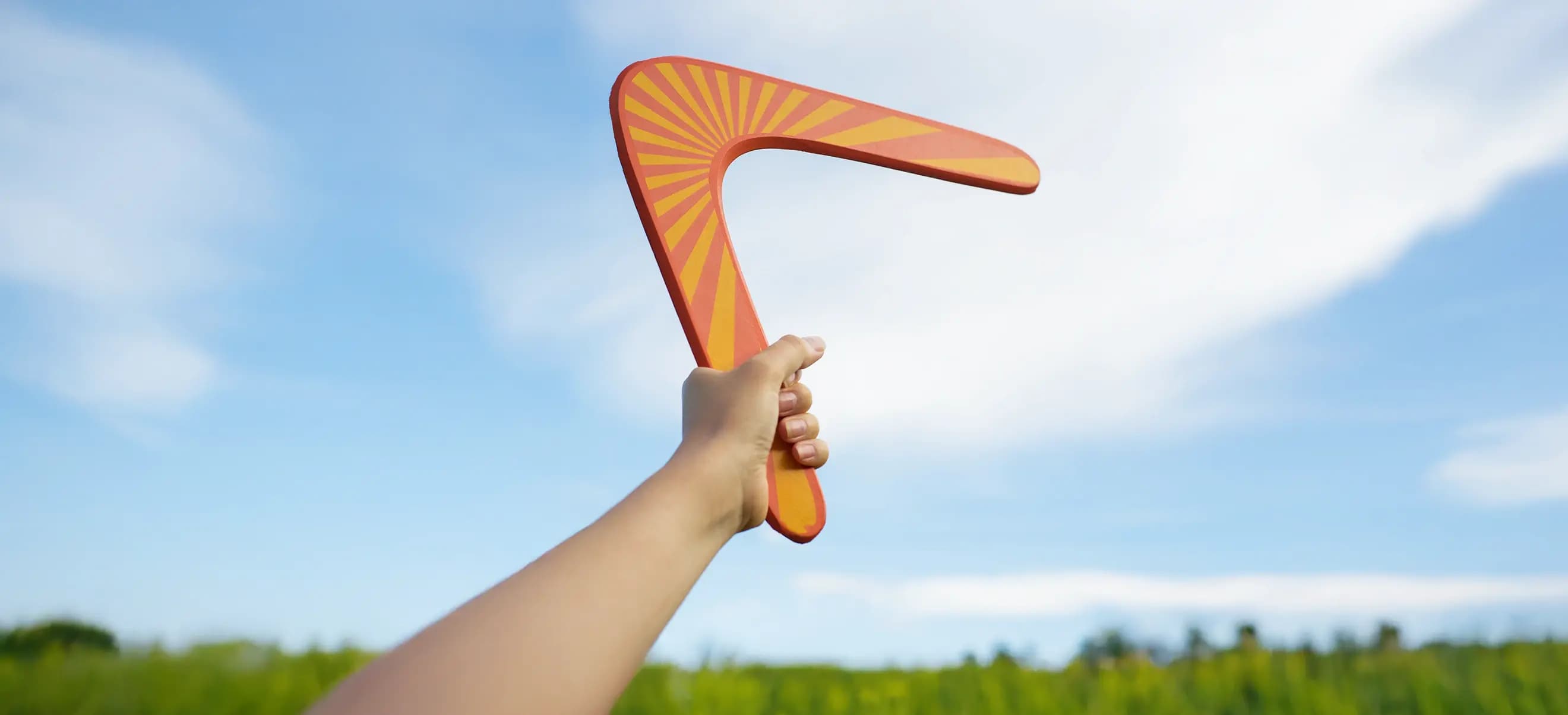 A hand holds an orange and yellow striped boomerang against a blue sky with clouds.